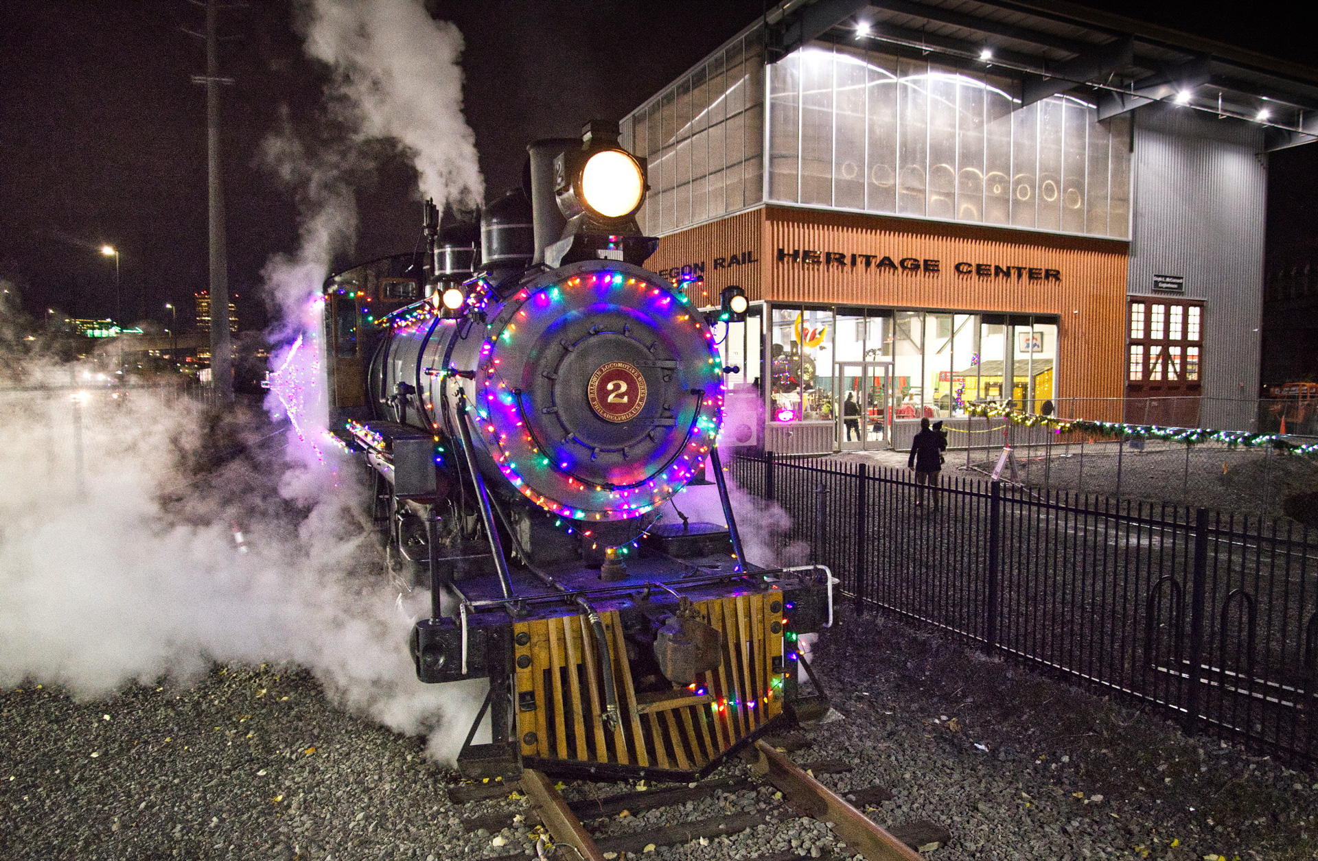 The Holiday Express train at night with festive lights on it and steam coming out of it.