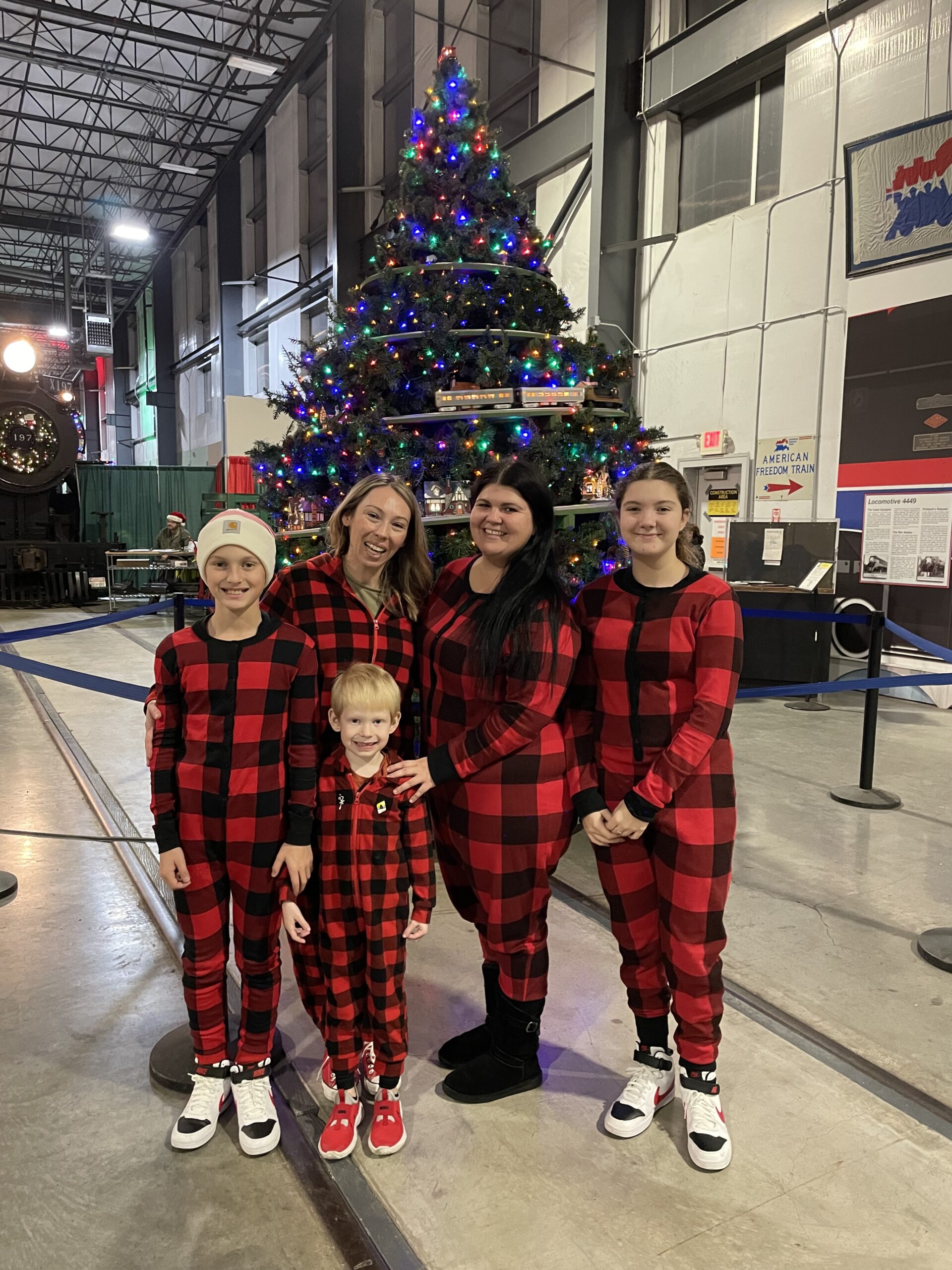 A family of 5 standing in front of a Christmas tree at the Oregon Rail Heritage Center in matching red plaid pajamas.