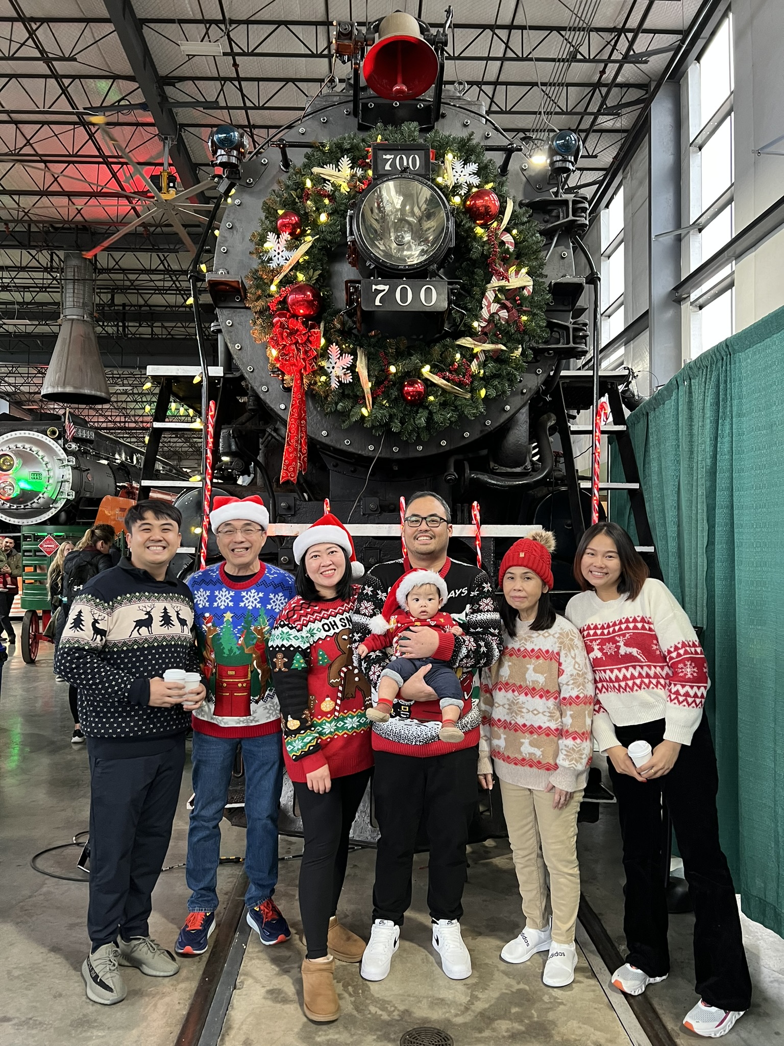 A family wearing Christmas sweaters posing in front of a steam locomotive decorated for the holidays.